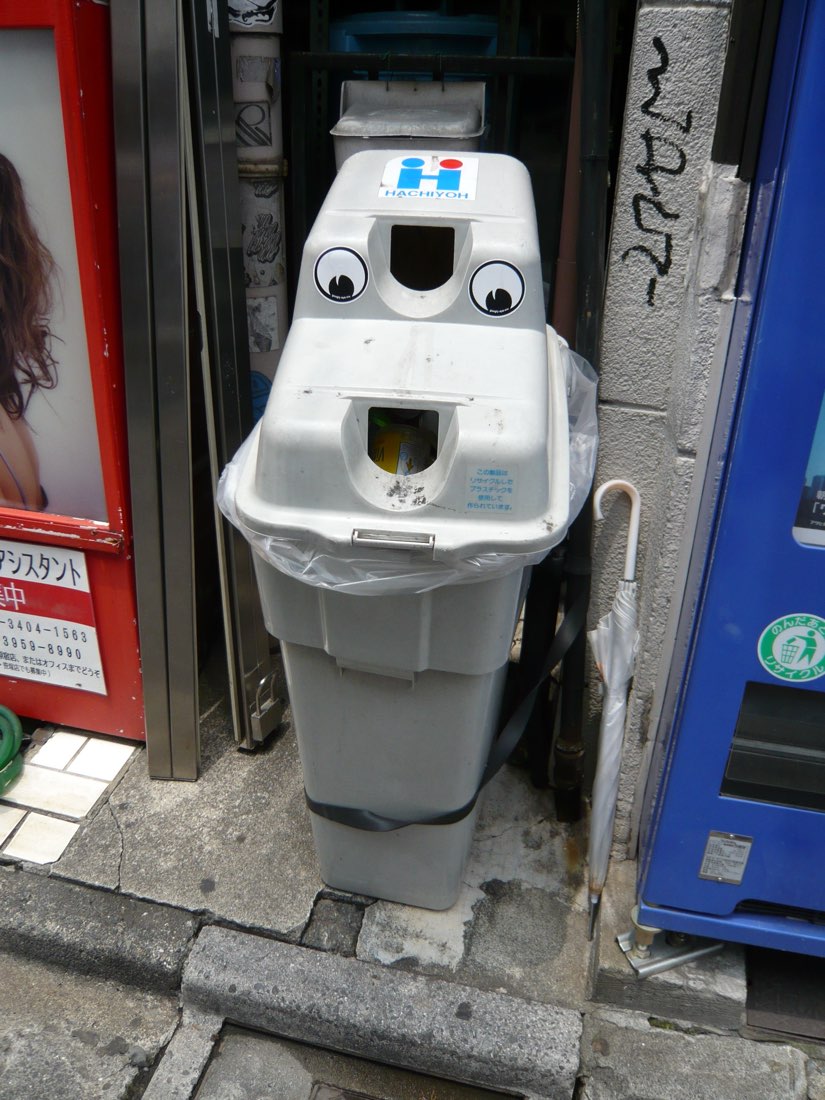 A grey street litter bin with two googly eyes stuck above its opening.
