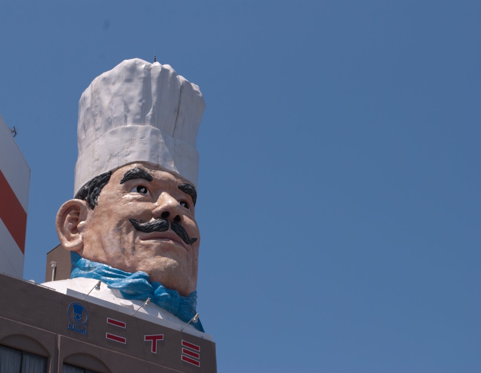 A giant chef statue with a tall white hat and curled moustache, photographed from below against a clear blue sky.