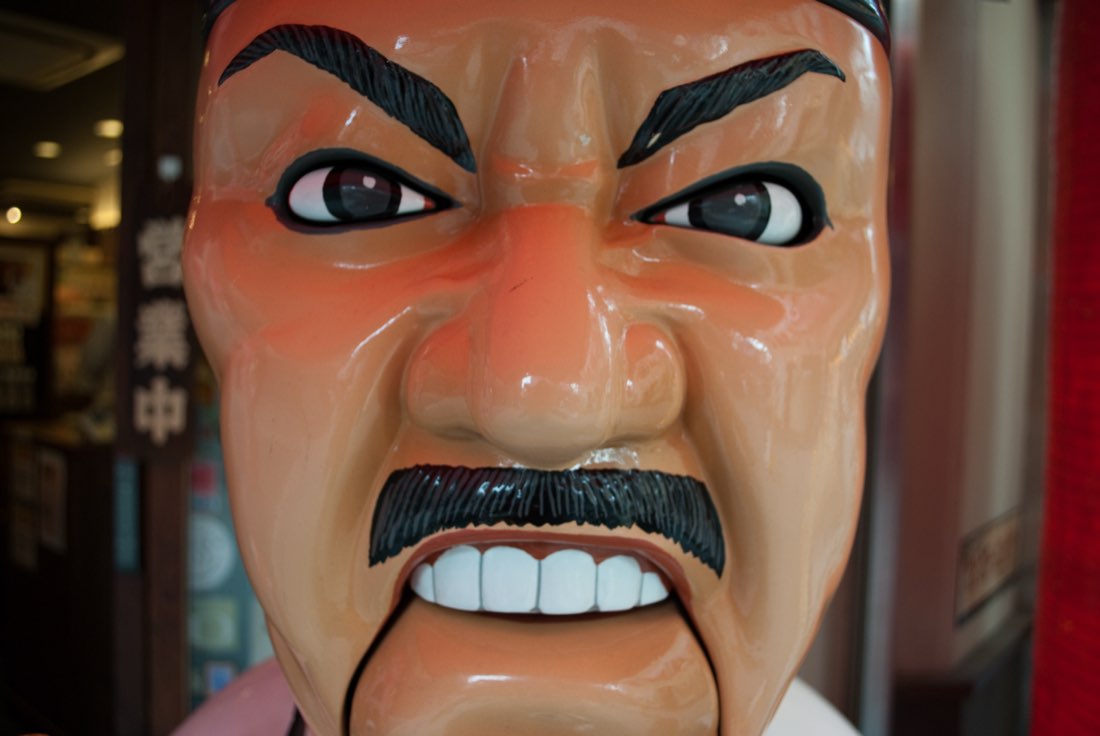 Close-up of Daruma Daijin, a scowling chef mascot with bulging eyes, thick eyebrows and a black moustache.