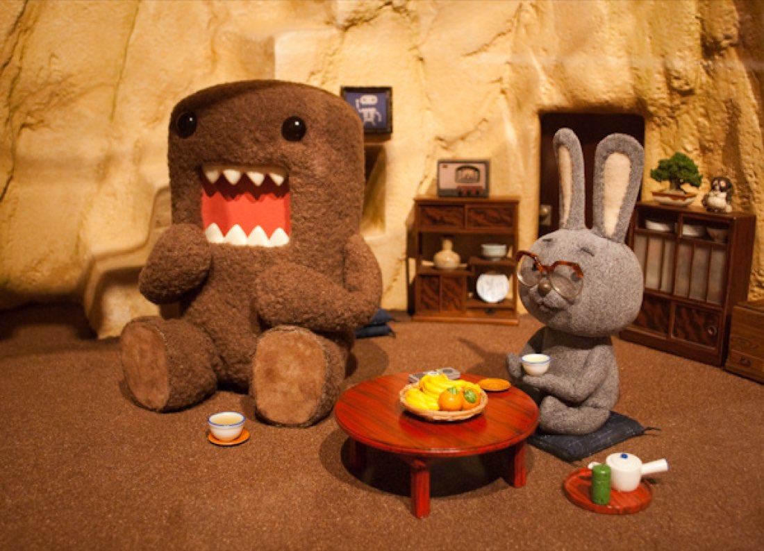 Domo-kun, a fuzzy brown rectangular creature with a wide toothy mouth, in a diorama tea room with a rabbit.