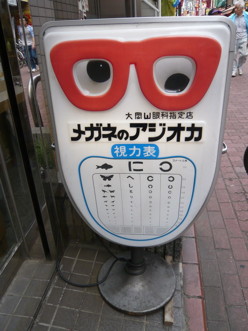 A giant pair of red spectacles mounted on a stand with an eye-test chart, outside an optician.