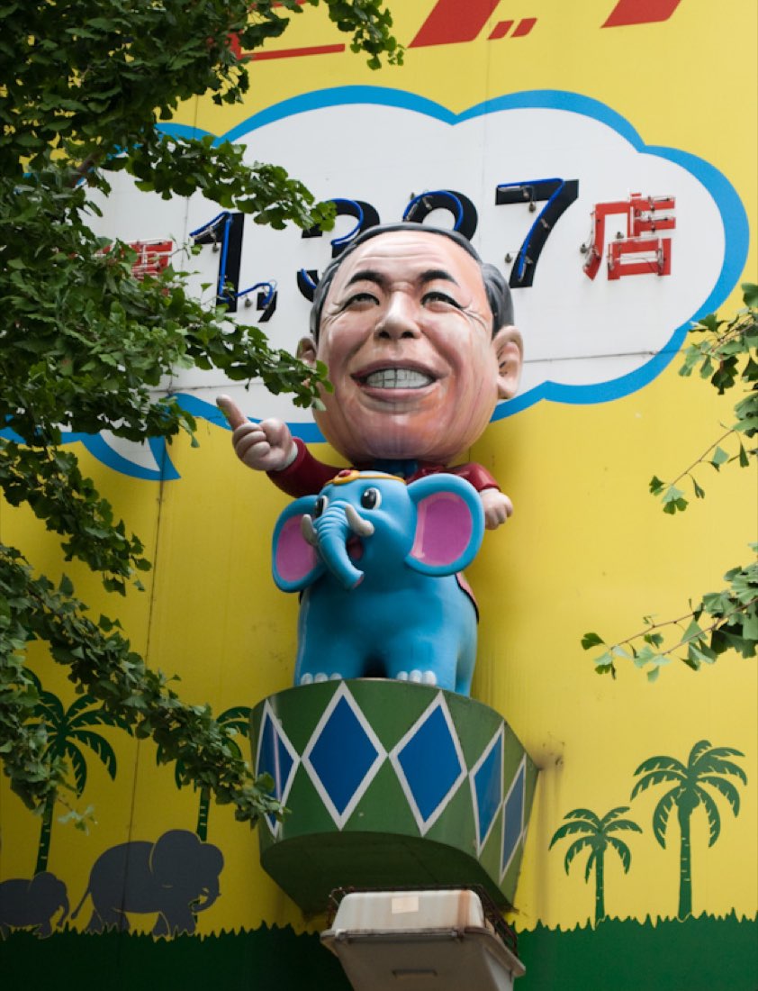 A grinning man in glasses riding a small blue elephant, on a pharmacy storefront sign reading 1,327 stores.