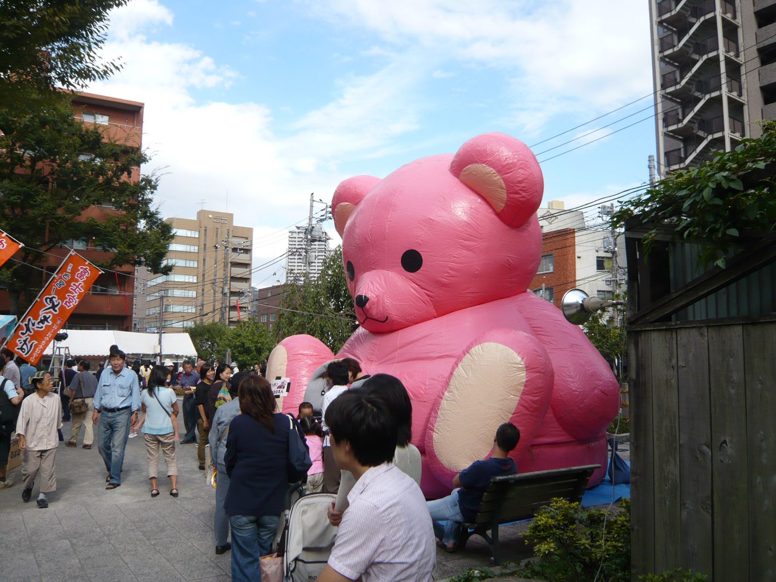 An enormous pink inflatable bear looming over a crowded festival street, trees in the foreground.