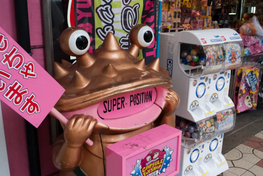 Kanegon, a bronze-coloured monster with a clam-shaped head and coin slot, beside pink capsule-toy machines.