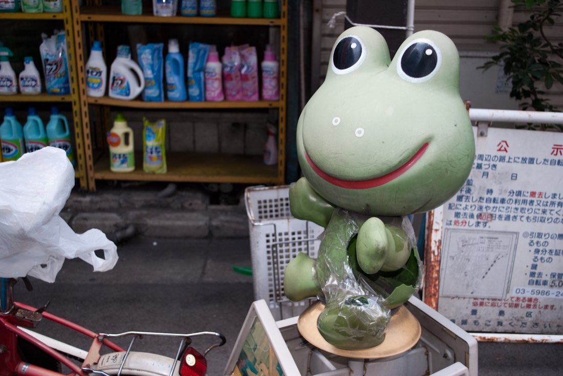 A worn green Kero-chan frog statue standing on a wooden stool outside a shop, arms out.