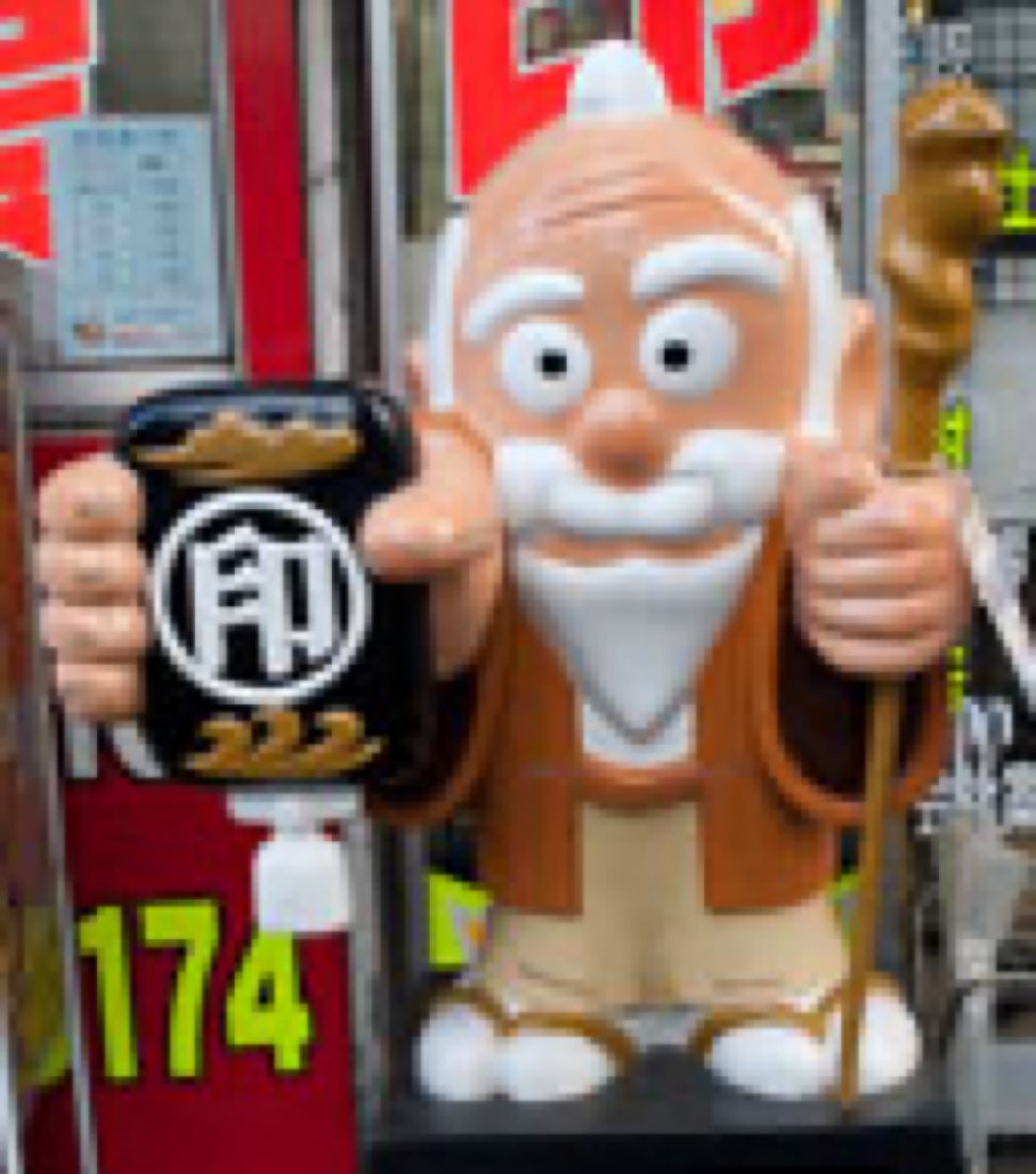 Koumon-sama, an elderly bearded mascot in traditional dress holding a seal case, outside a seal shop.