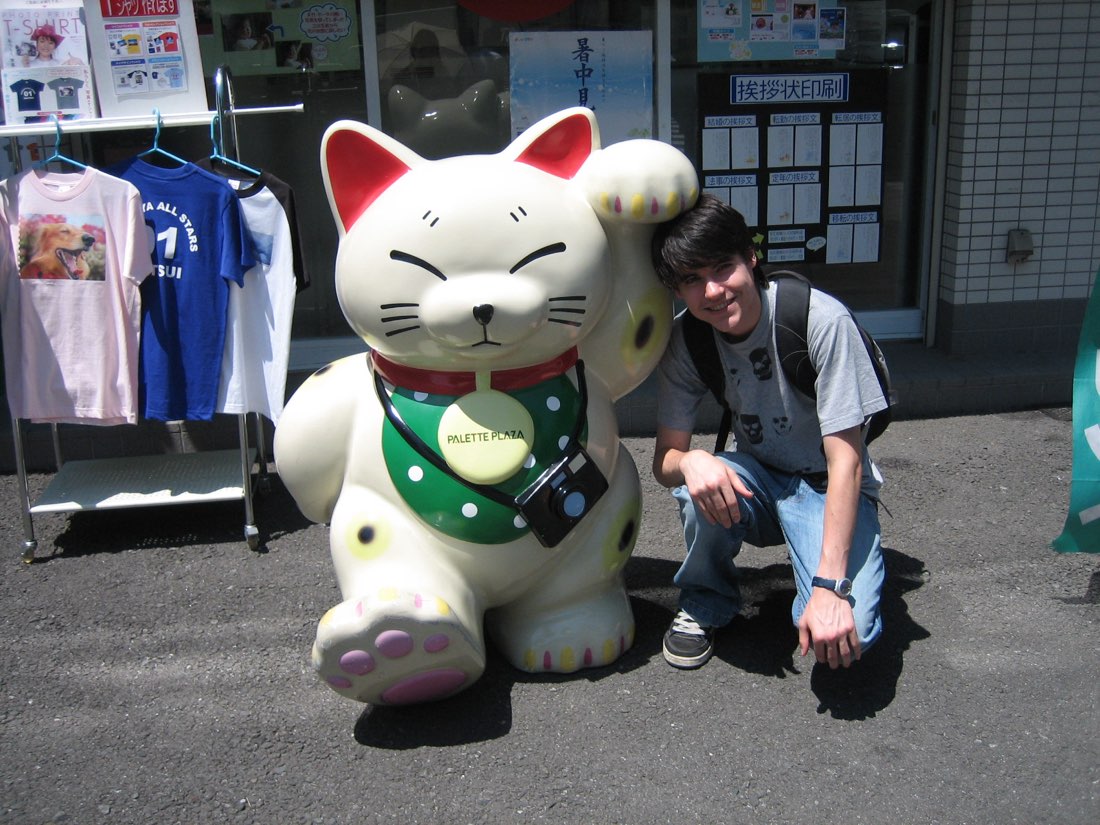 A person crouching beside a maneki-neko statue roughly twice their size, outside a shop.
