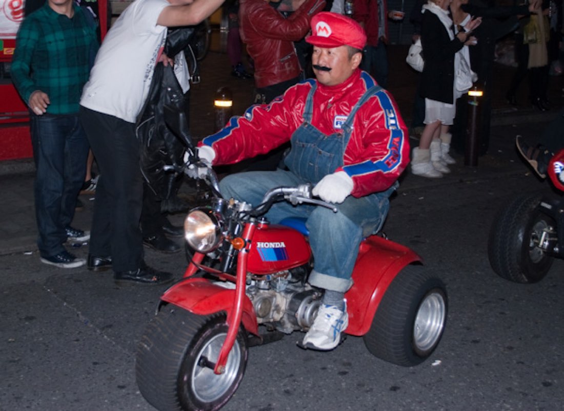 A man dressed as Mario riding a very small red motor-trike through a city street at night.