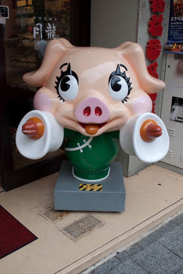 A pig mascot with long eyelashes holding up two plates outside a restaurant.