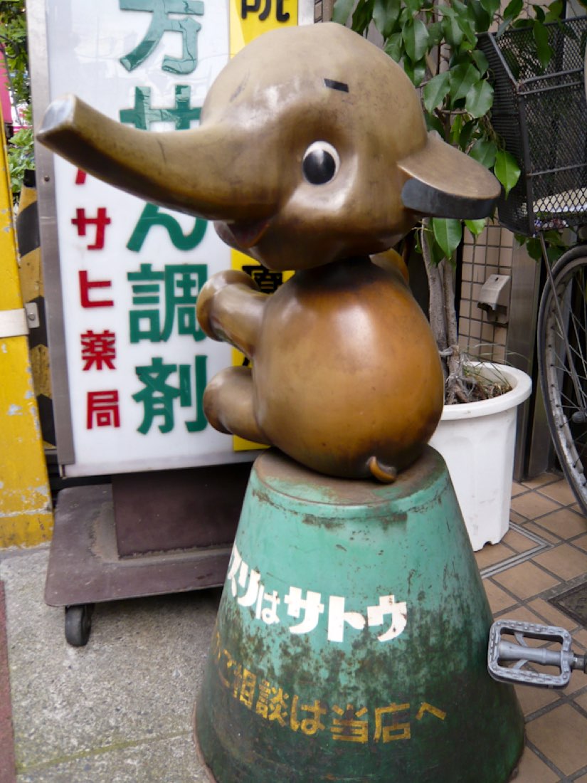 A weathered bronze-coloured Sato elephant on a worn green stand marked Asahi.