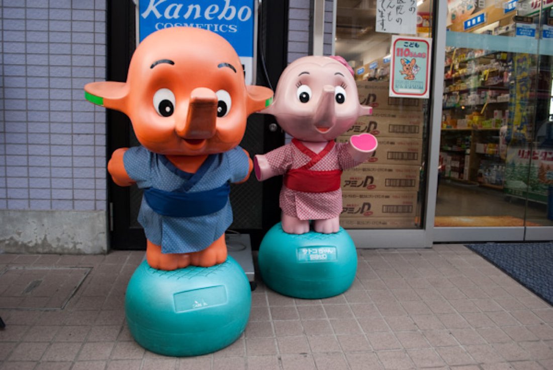 Satoko-chan, a pink elephant mascot, dressed in a red patterned kimono outside a pharmacy.