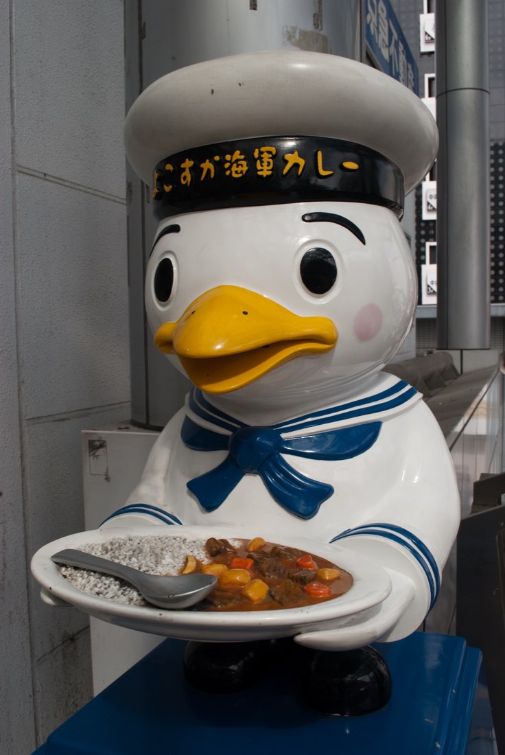 Sukare-chan, a large seagull mascot in a navy sailor uniform, holding a plate of curry and rice with a spoon.