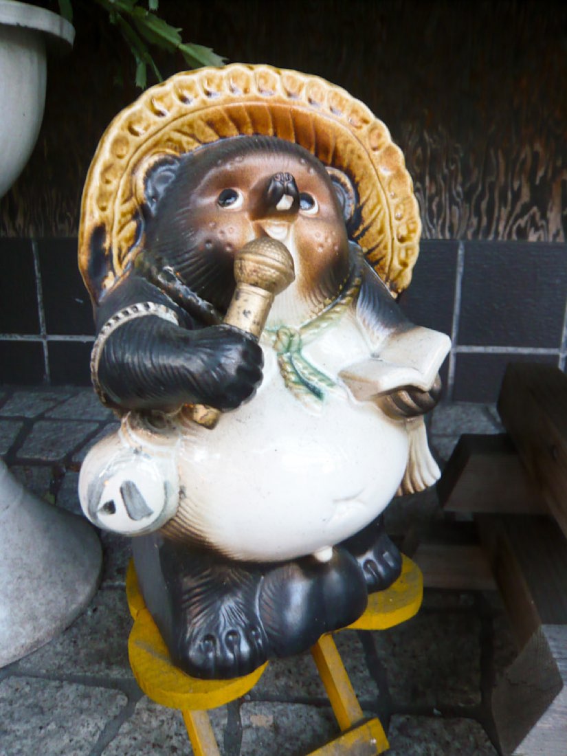 A ceramic tanuki statue in a straw hat holding a microphone, standing on a doorstep.