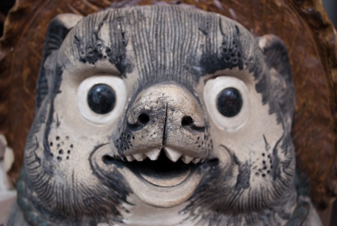 A dark, weather-worn tanuki statue with pale bulging eyes and a toothy open mouth.