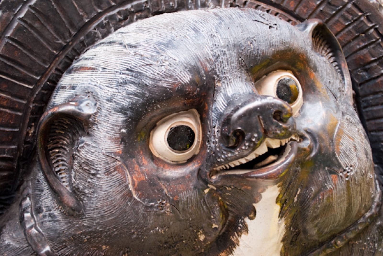 Extreme close-up of a weathered wooden tanuki face, eyes wide, whiskers scored into the grain.