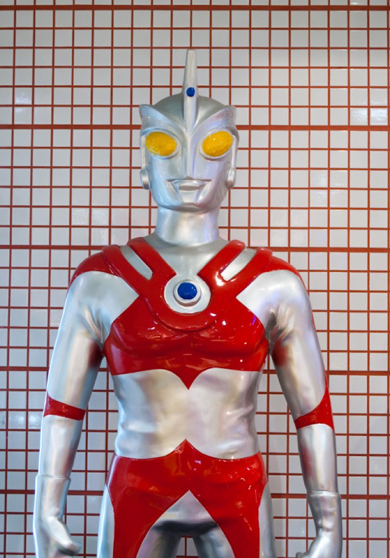 An Ultraman statue in silver and red, hands on hips, reflected shopfronts behind.