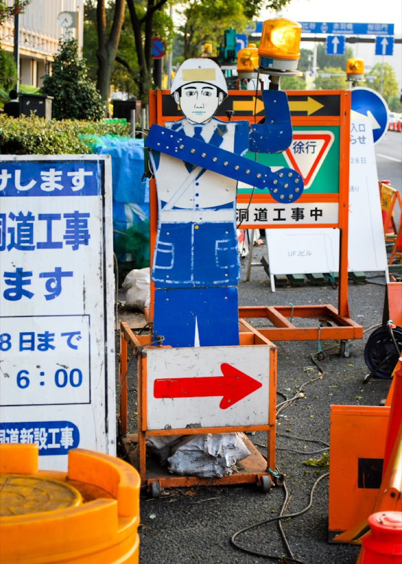 A flat wooden workman figure in blue overalls beside roadworks, next to a large arrow sign.