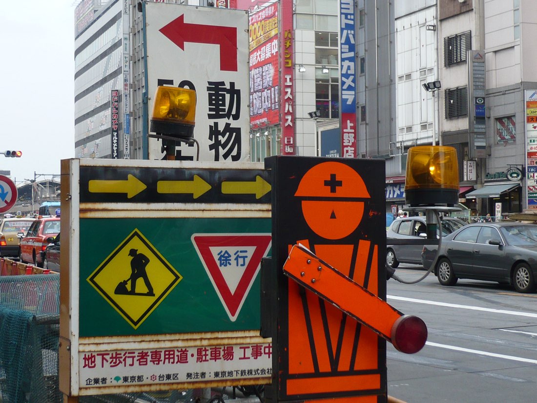A cartoon hard-hatted workman painted on a street sign, pointing left, among other traffic signs.