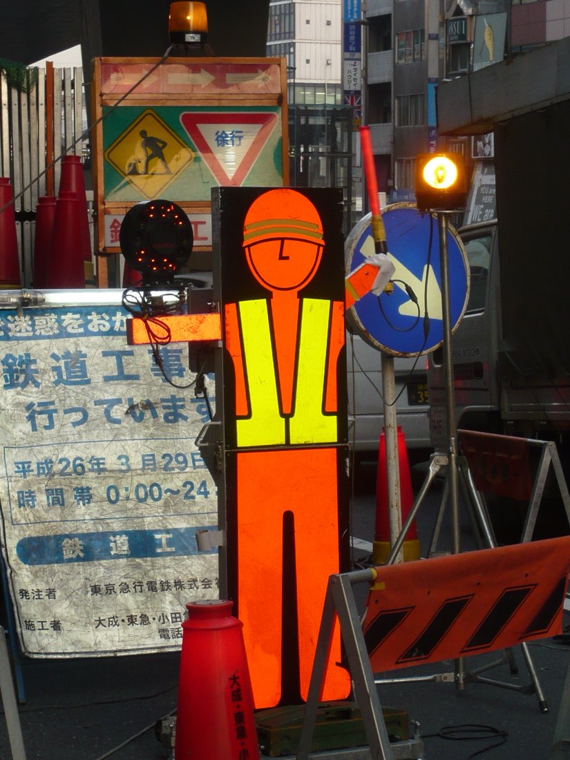 Two flat plywood workmen in day-glo orange vests standing guard at a rail-work barrier.